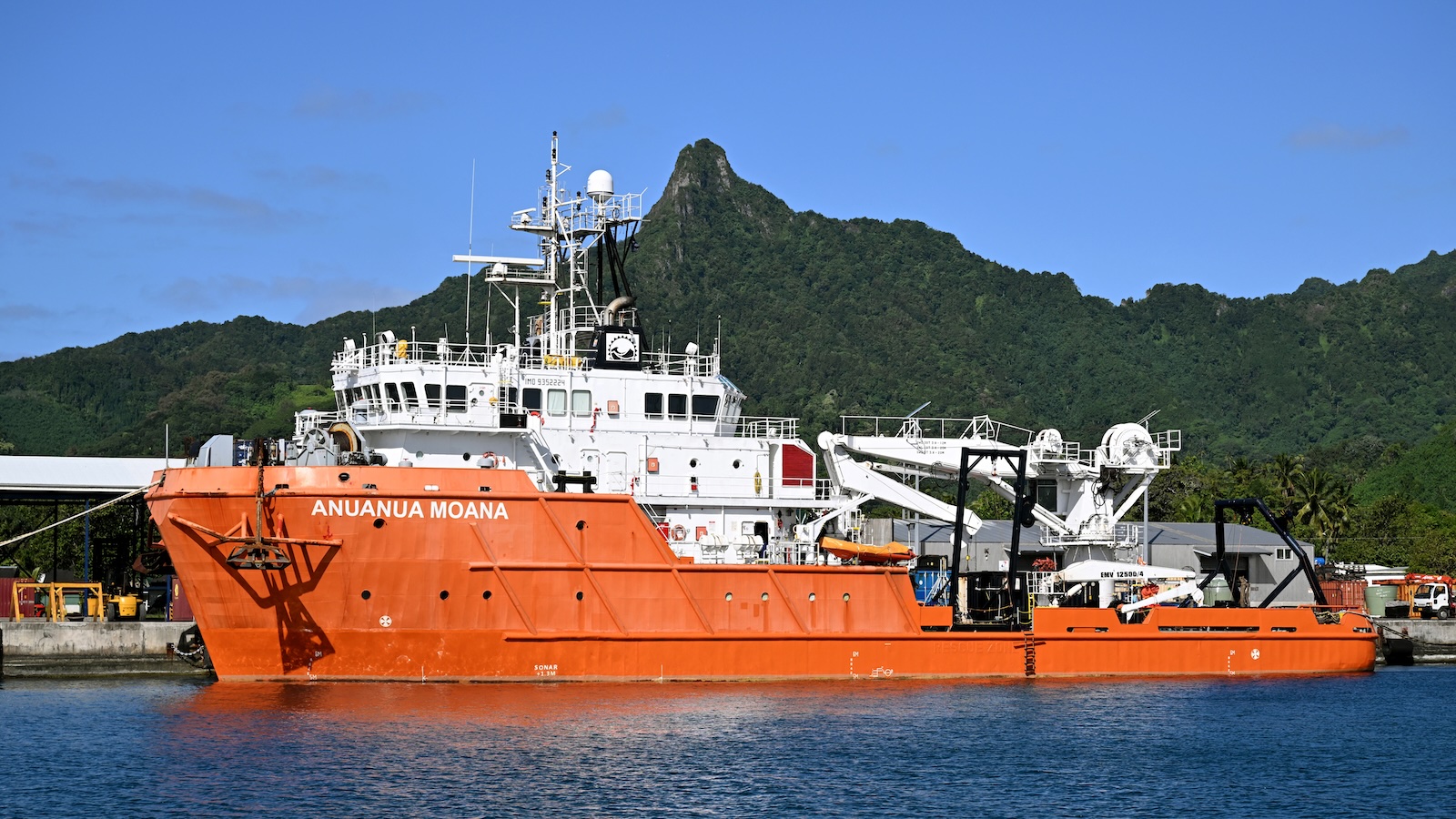 an orange ship in front of a forested tropical island sihoette sits in front of a port area. its hull says Anuanua Moana in white paint and capital letters.