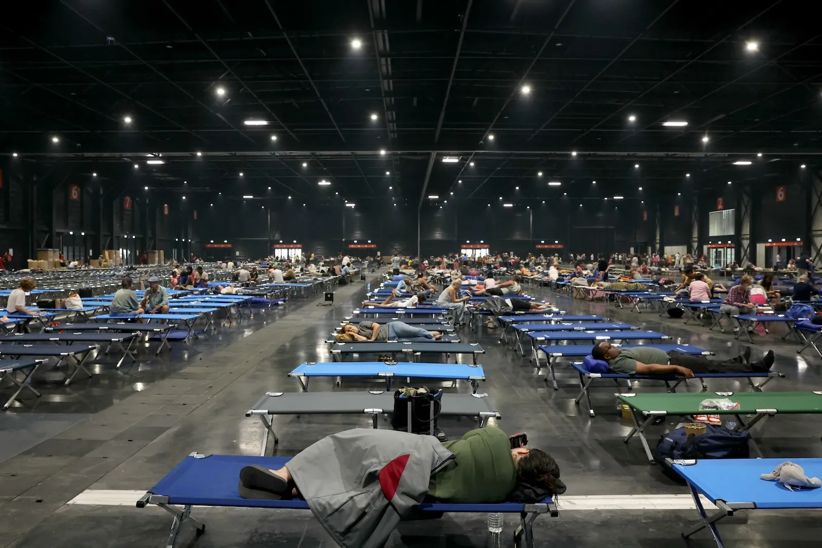 several rows of blue and grey cots, with dozens per row, strech into a large auditorium space. dozens of people are visible sitting, lounging, or sleeping on them.