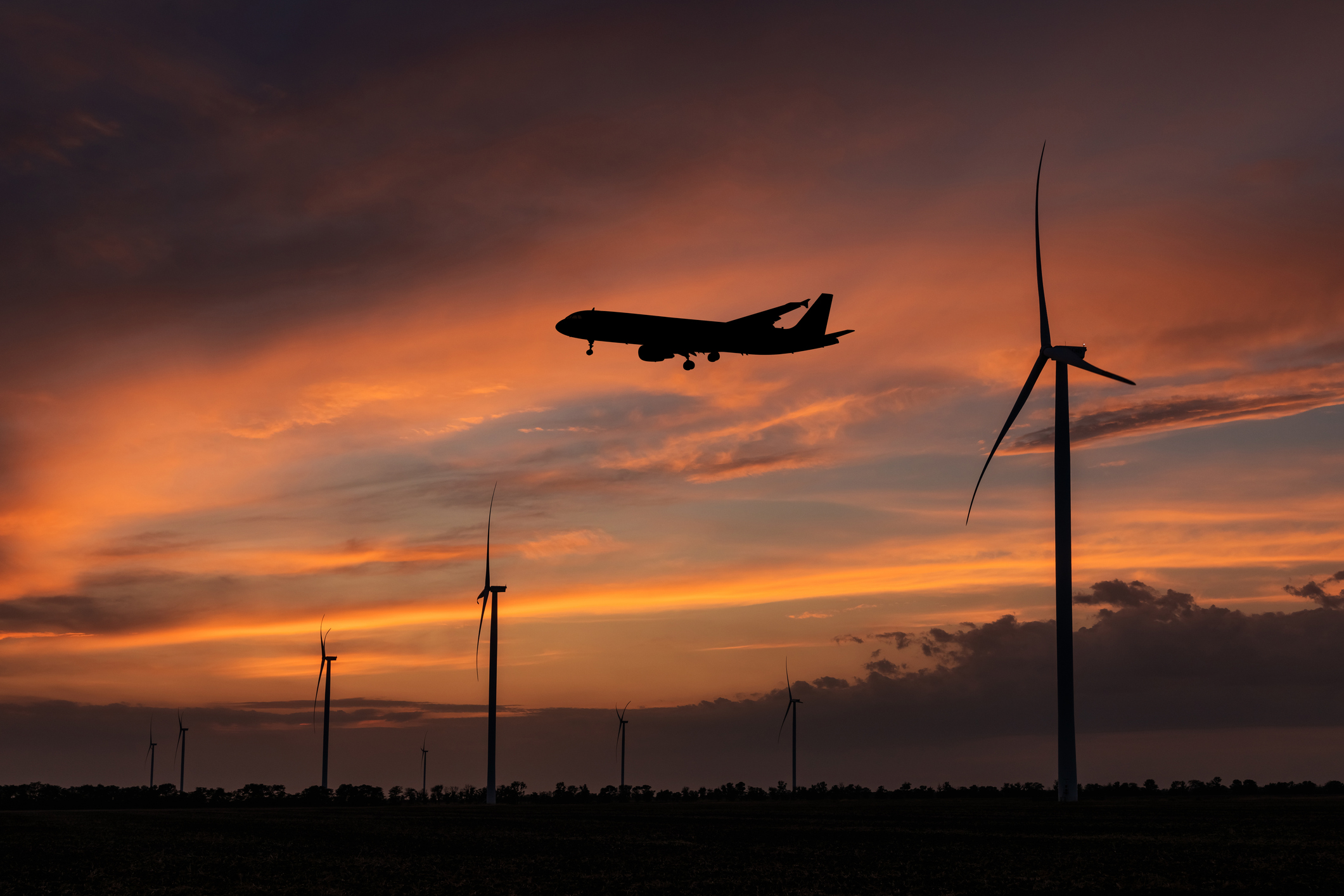 An airliner flies over a line of wind turbines at sunset.