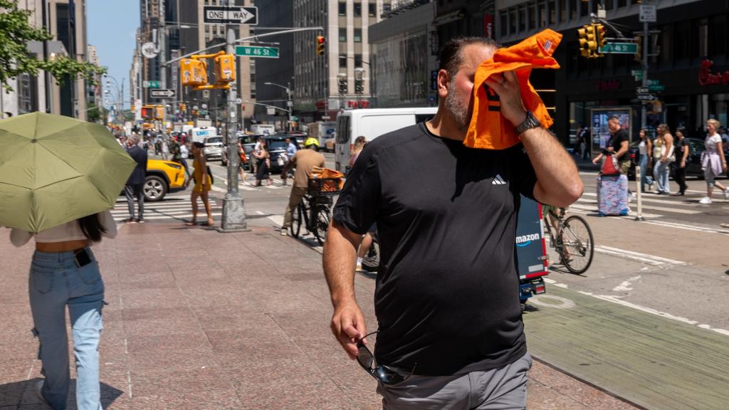 a man wearing a black shirt walking down the street in new york city's midtown wipes the sweat off his brow with a small orange towel