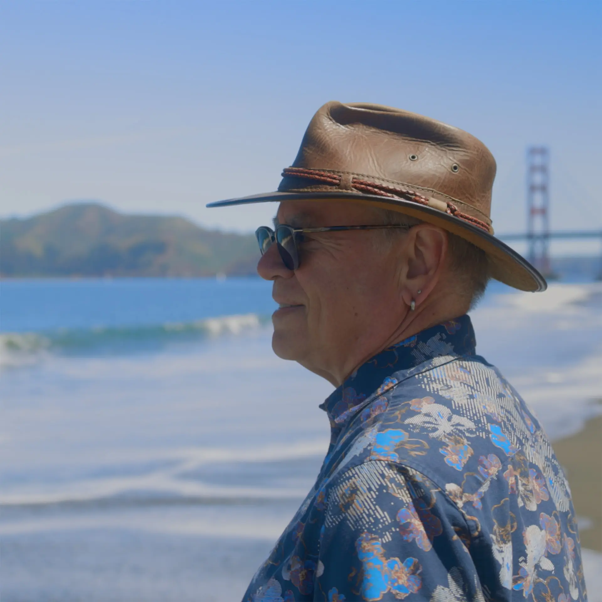 A man in a hat looks off screen at the ocean. The Golden Gate Bridge is visible in the background.