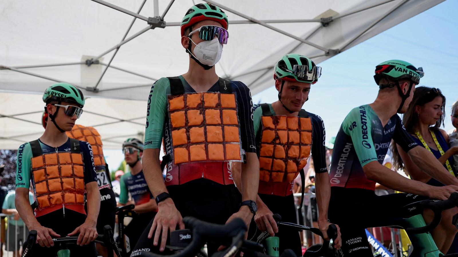 Cyclists competing in the Tour de France cool themselves with ice vests as they stand in the shade of an awning.