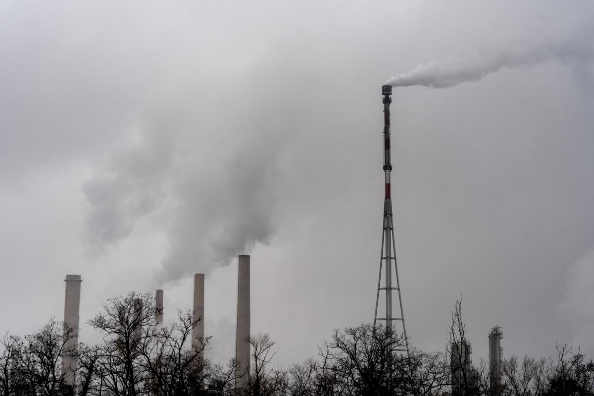 Black-and-white photo of the chimneys at a refinery emitting thick white smoke under a cloudy sky