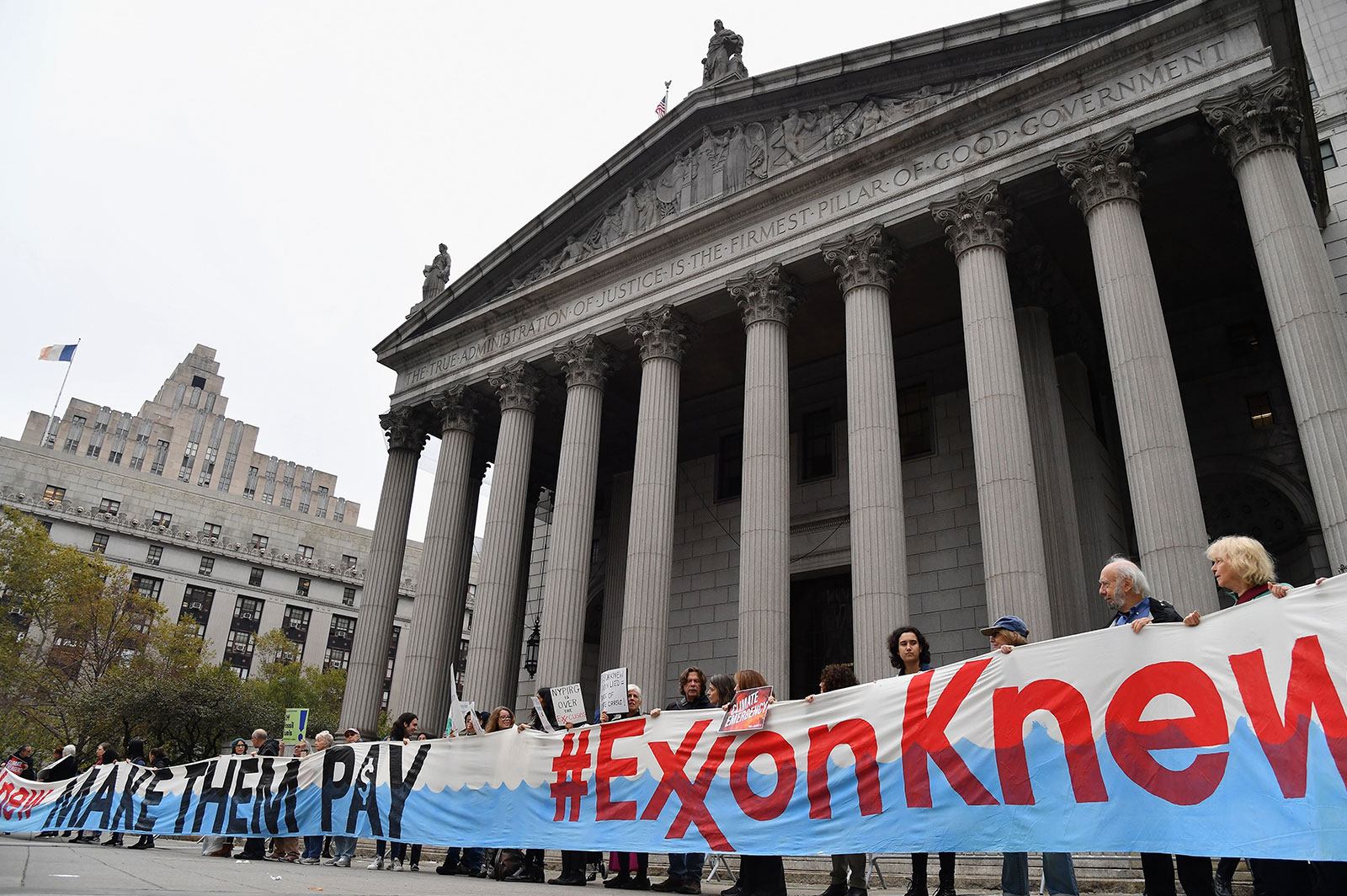 people stand in front of the New York Supreme Court building holding a large banner that says '#ExxonKnew'