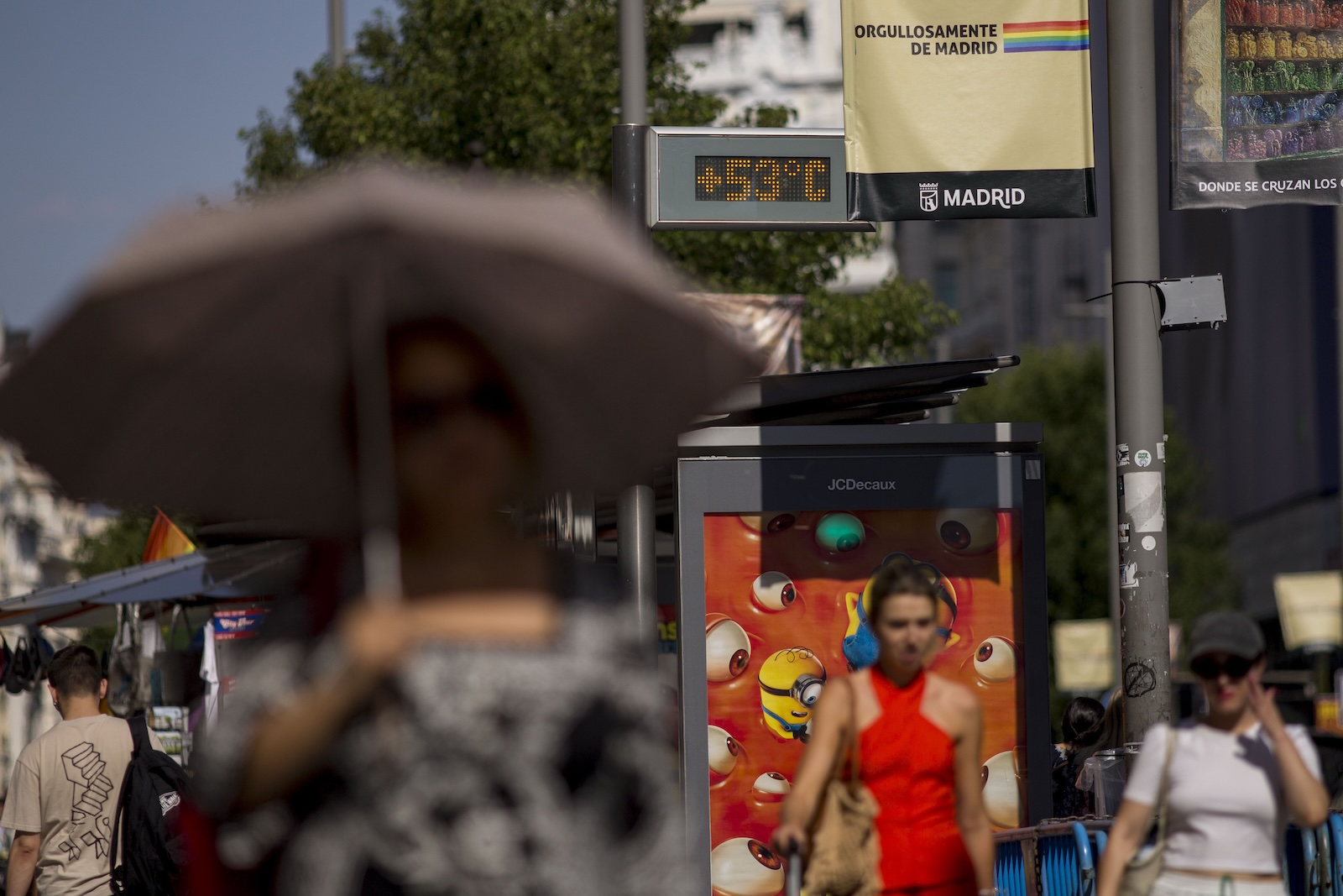 people under umbrellas walk through a seemingly busy area passing a busstop to their left with a thermometer screen that reads +53 degrees C