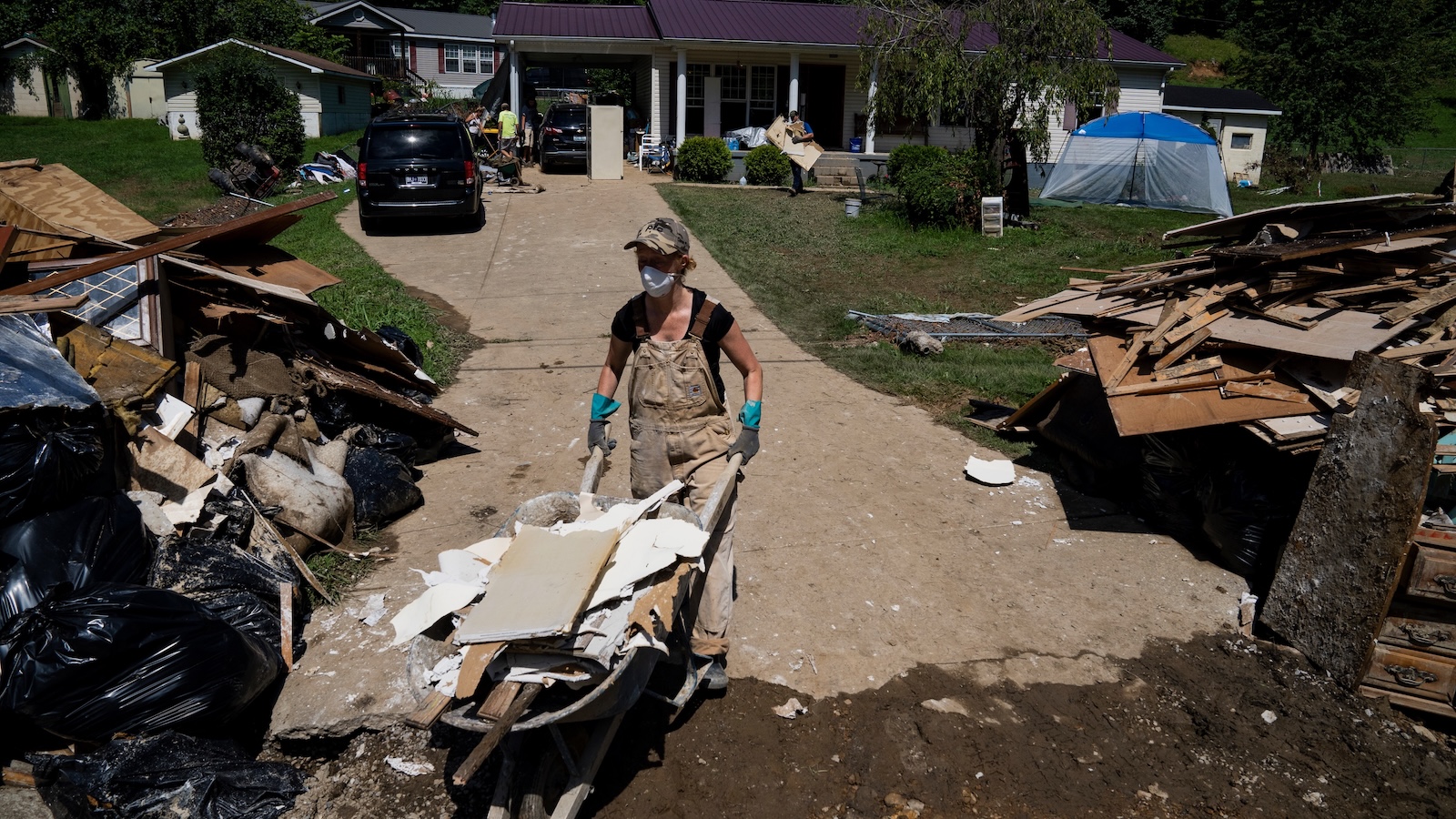 A woman wearing a mask and coveralls pushes a wheelbarrow full of debris down the debris-strewn driveway of her flood-damaged home.