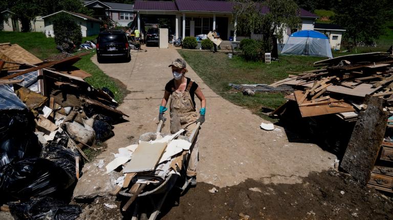 A woman wearing a mask and coveralls pushes a wheelbarrow full of debris down the debris-strewn driveway of her flood-damaged home.