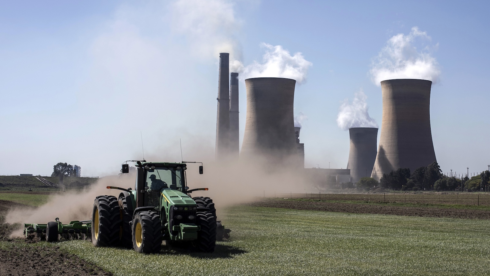 A tractor drives along a farm, with a large coal-fired power plant in the background