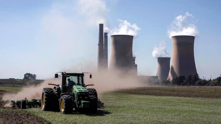 A tractor drives along a farm, with a large coal-fired power plant in the background
