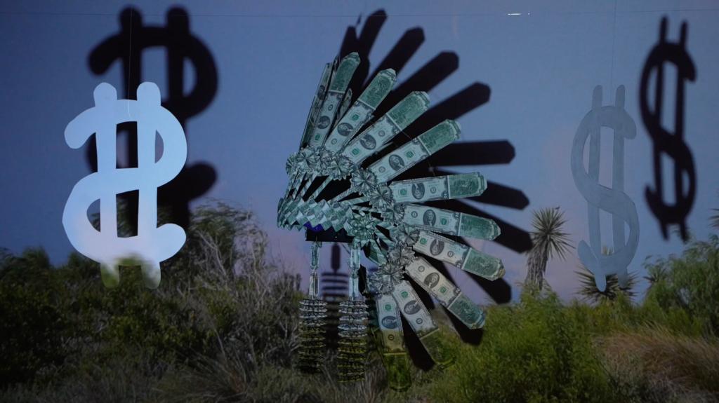 A diarama consisting of an iconic Indigenous headdress made of $2 bills hanging in front of a screen next to cut out dollar bills. Over the tableau, a video of a Texas SpaceX launch plays with the land in the forefront of the scene.