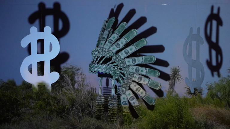 A diarama consisting of an iconic Indigenous headdress made of $2 bills hanging in front of a screen next to cut out dollar bills. Over the tableau, a video of a Texas SpaceX launch plays with the land in the forefront of the scene.