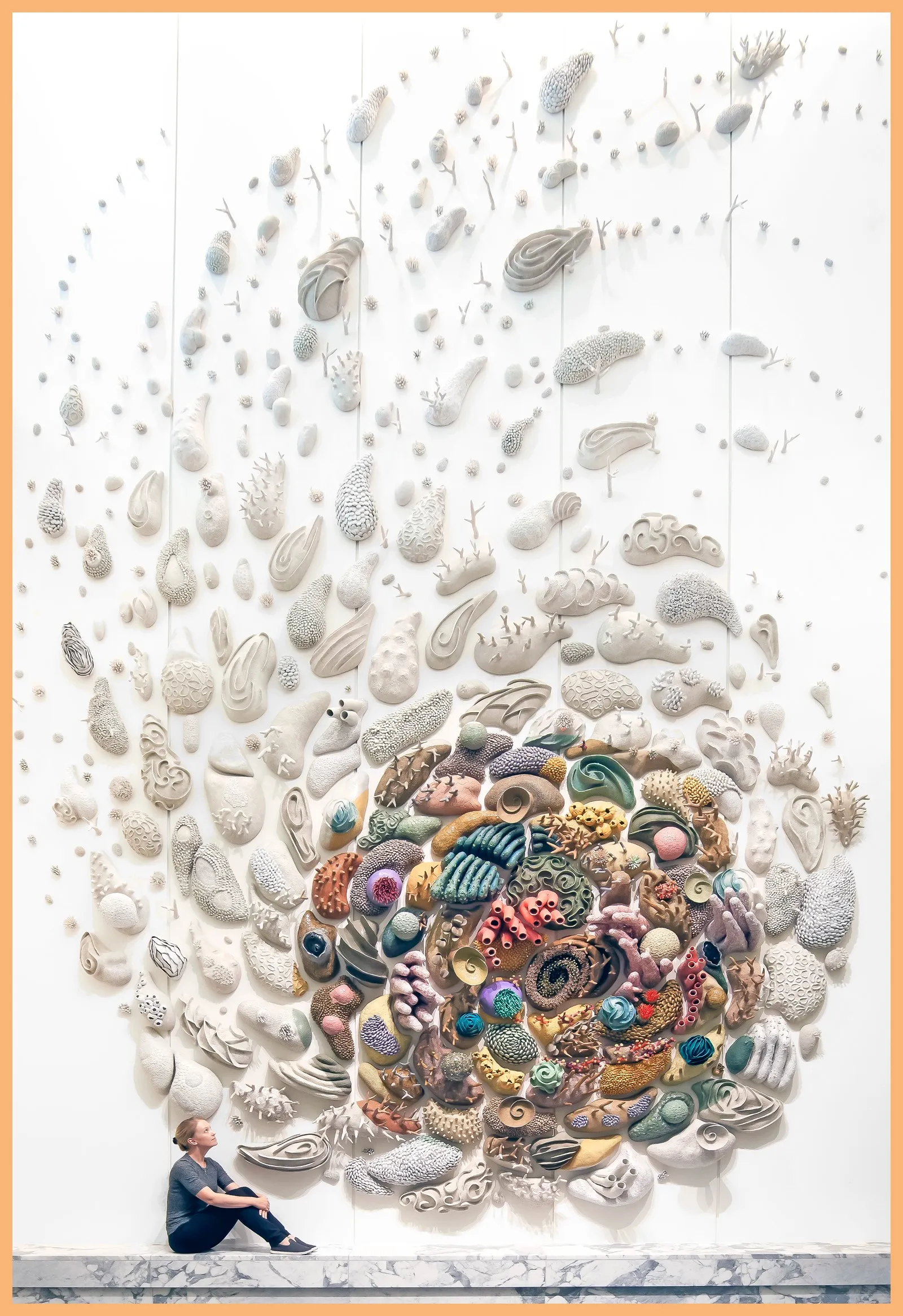 A photo shows a large ceramic sculpture, resembling a coral reef, mounted on a wall, with a woman sitting next to it for scale
