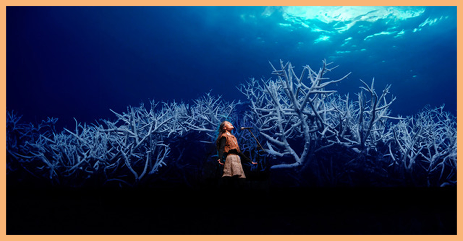 A woman stands before a microphone singing, with a screen displaying larger-than-life corals behind her