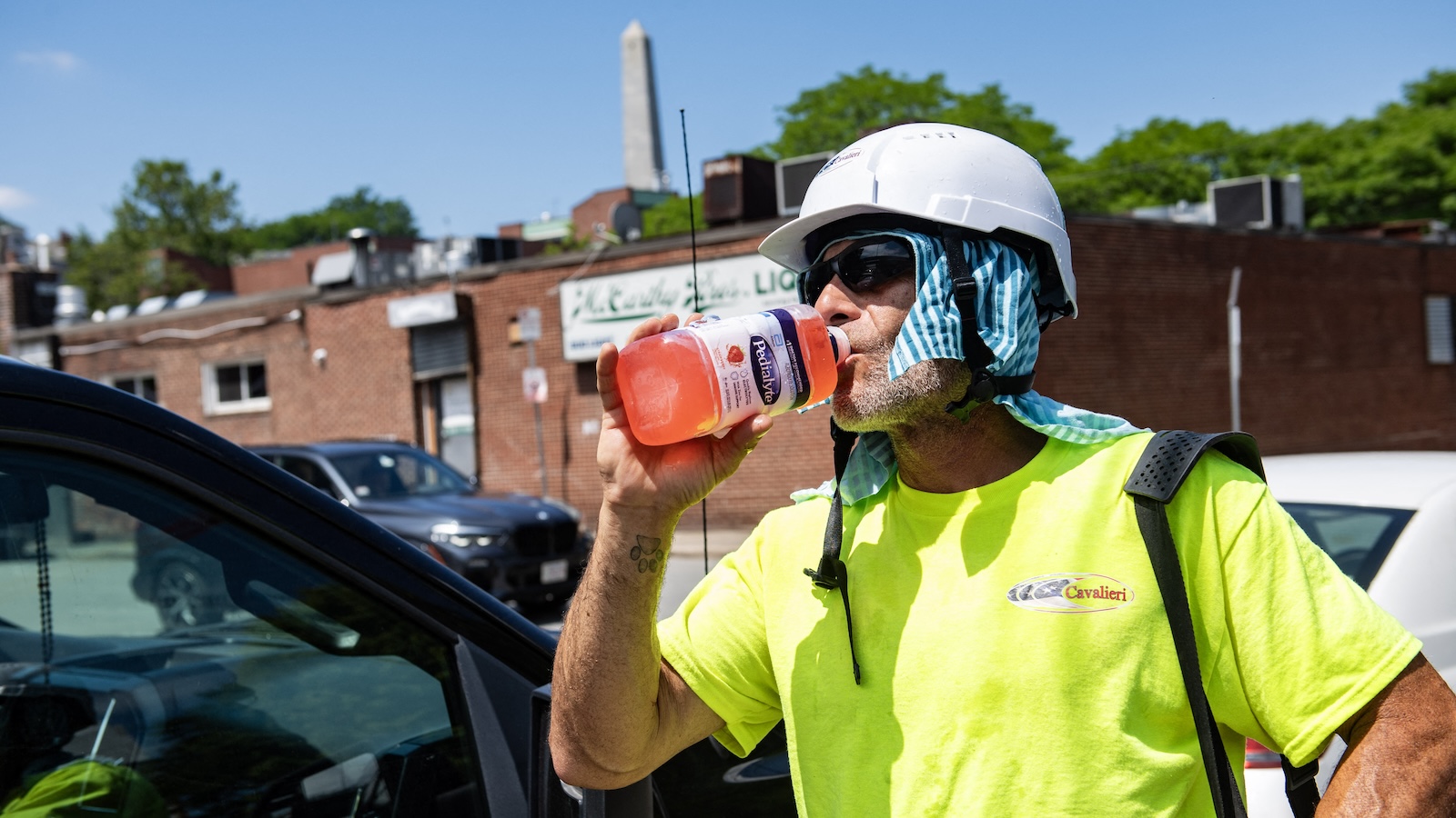 A construction worker in a bright yellow shirt wearing a white work helmet with a towel underneath takes a drink of an electrolyte beverage in the oppressive summer heat.