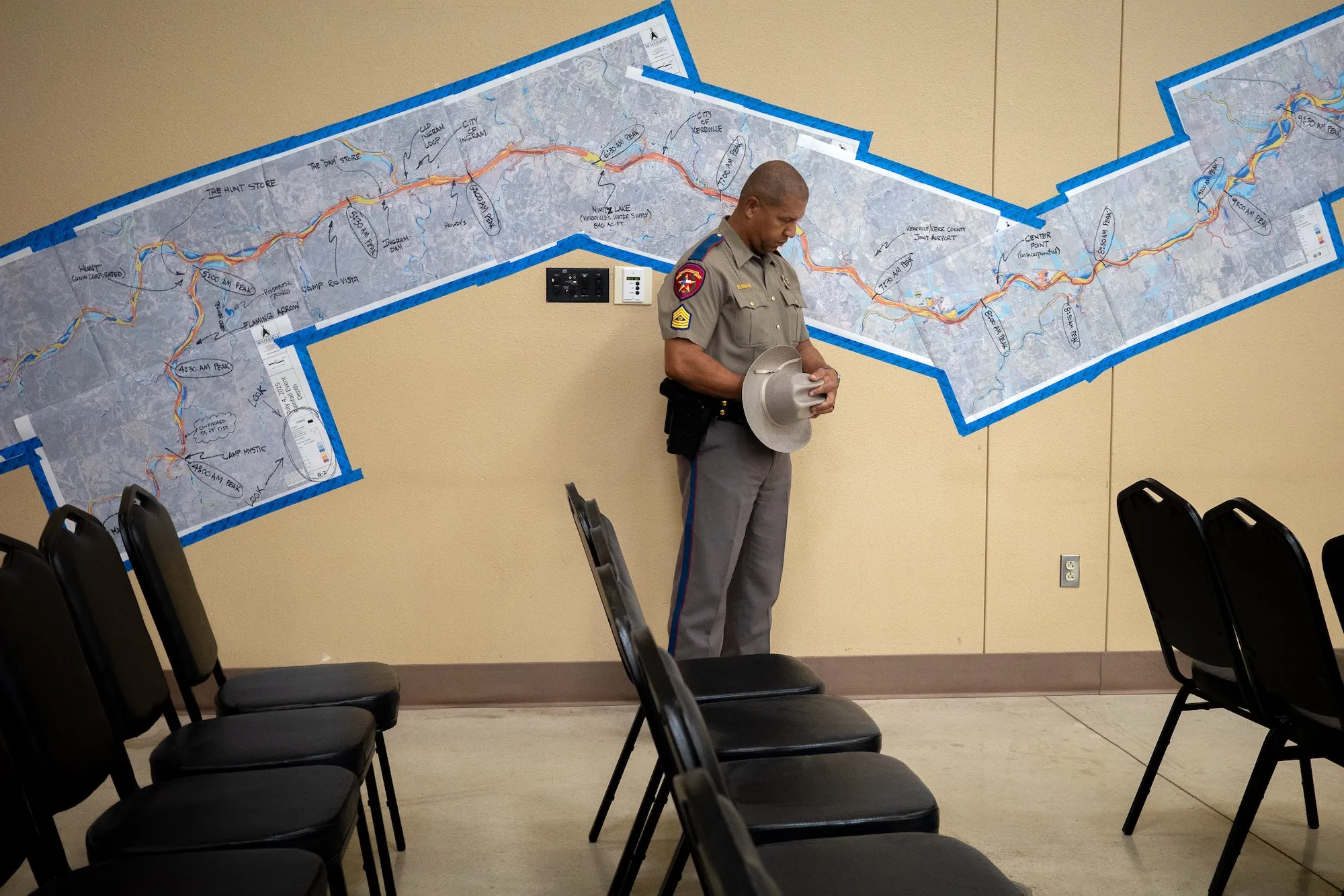 A man in a khaki uniform bends his head and holds his hat at waist height in front of a flood map pasted on a wall in a meeting room