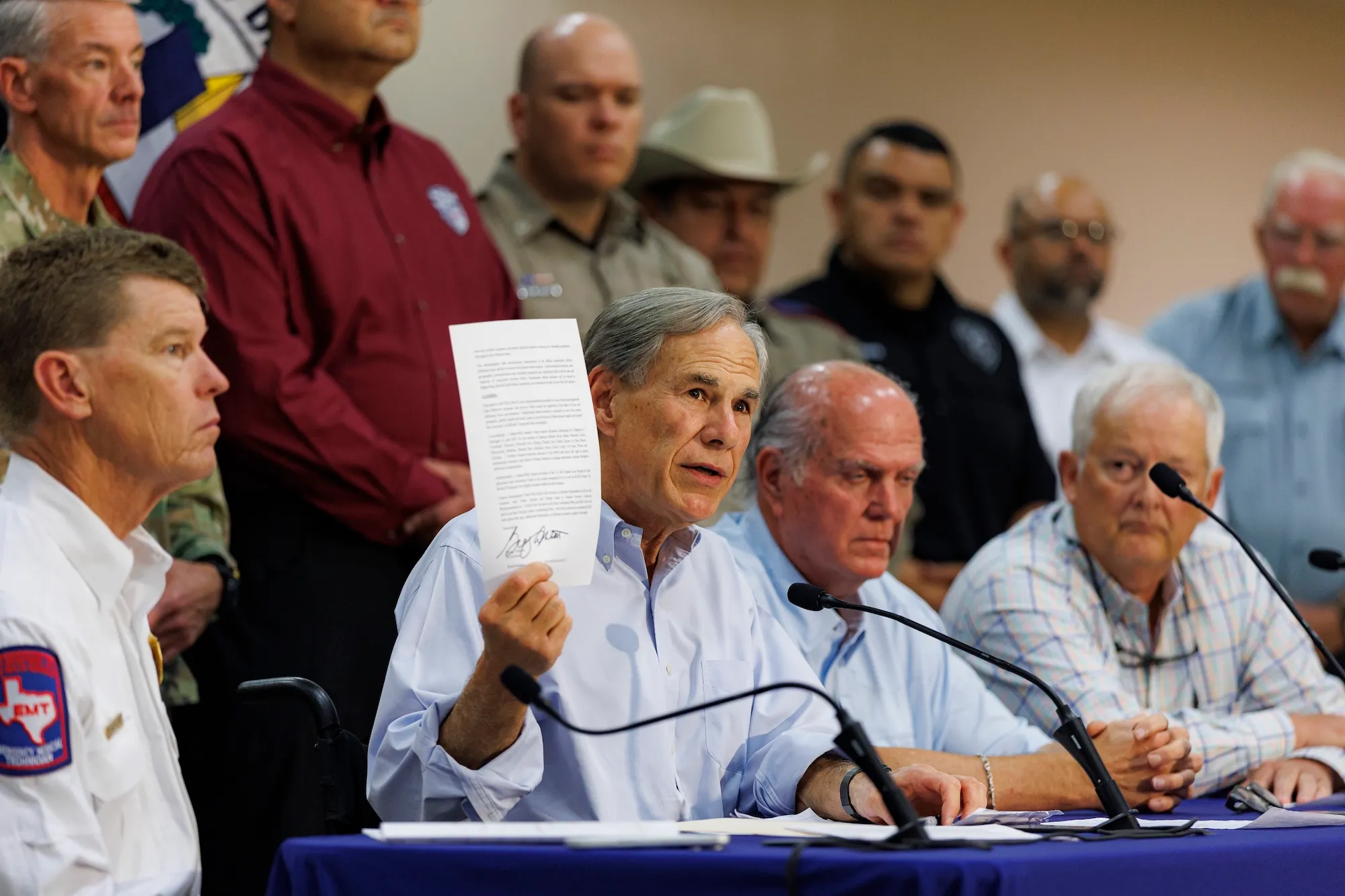 a man holds a piece of paper during a press conference with other people sitting and standing near him