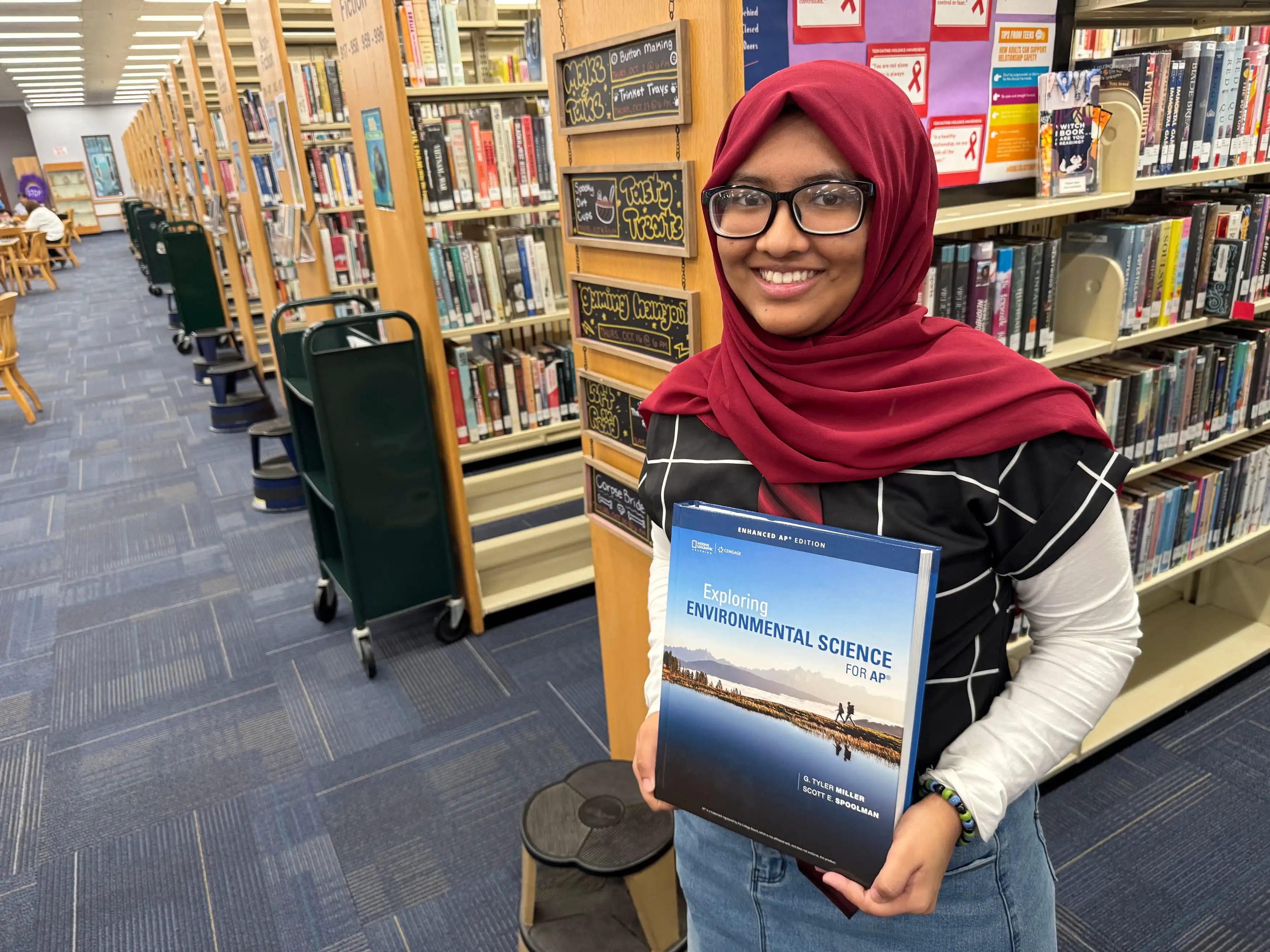 A girl wearing glasses holds a book up in a library