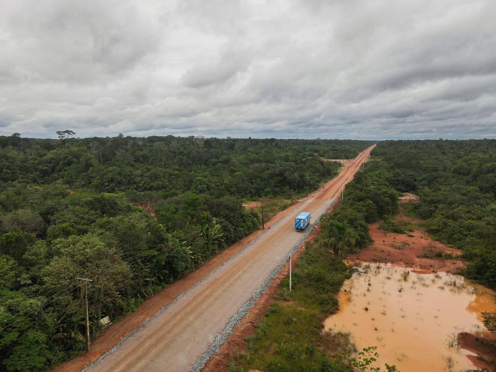 A blue truck drives along an unpaved section of a road cutting through the Amazon rainforest