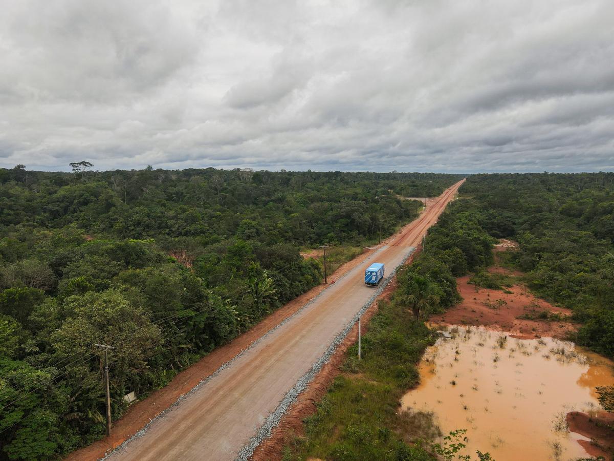A blue truck drives along an unpaved section of a road cutting through the Amazon rainforest