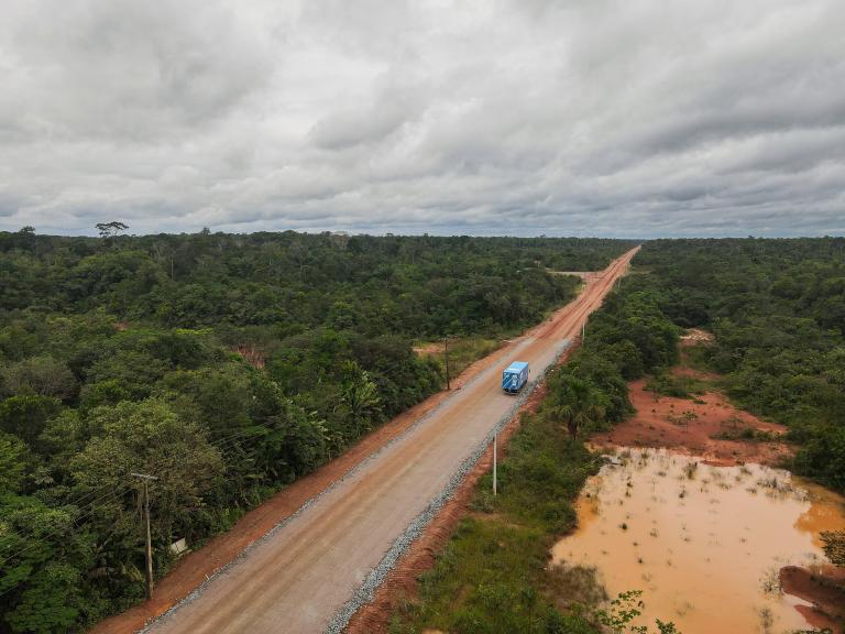 A blue truck drives along an unpaved section of a road cutting through the Amazon rainforest