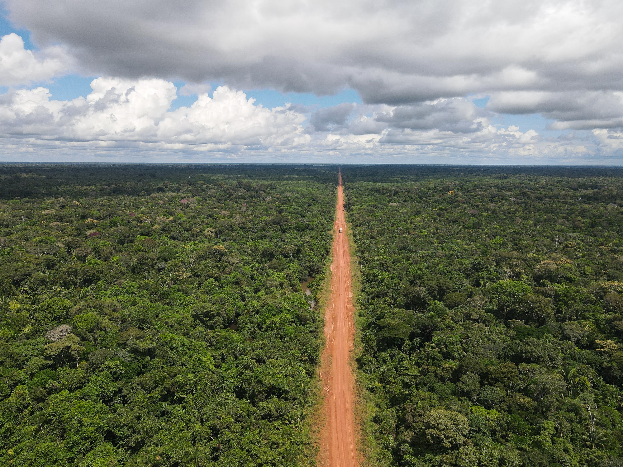 A blue truck drives along an unpaved section of a road cutting through the Amazon rainforest