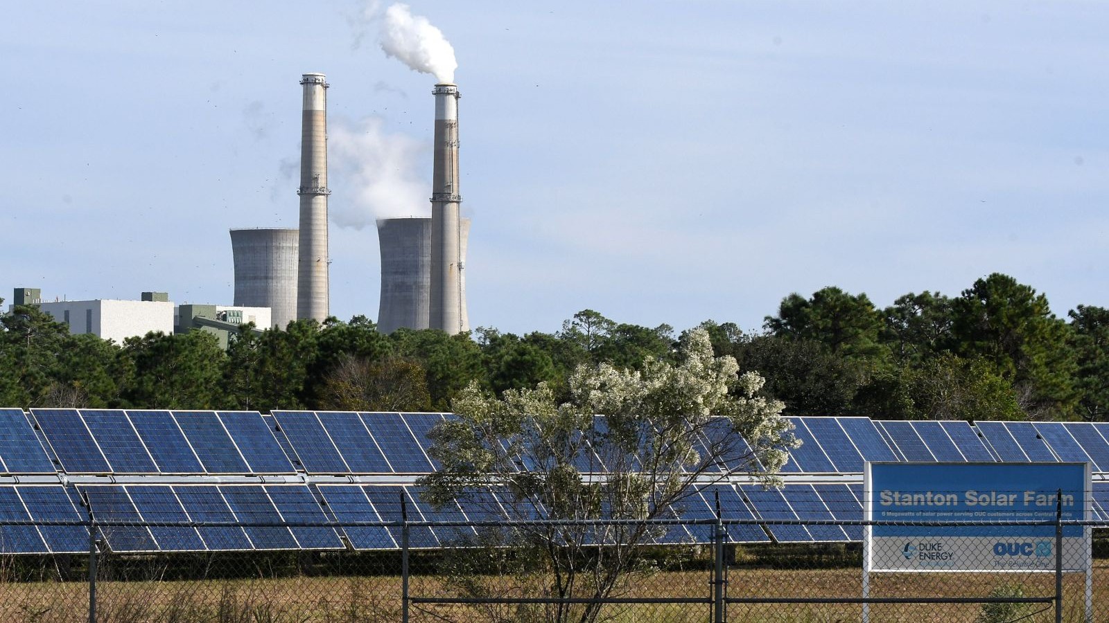 A solar farm takes in sun while smokestacks from a power plant puff in the background.