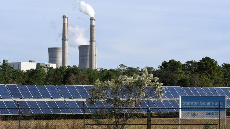A solar farm takes in sun while smokestacks from a power plant puff in the background.
