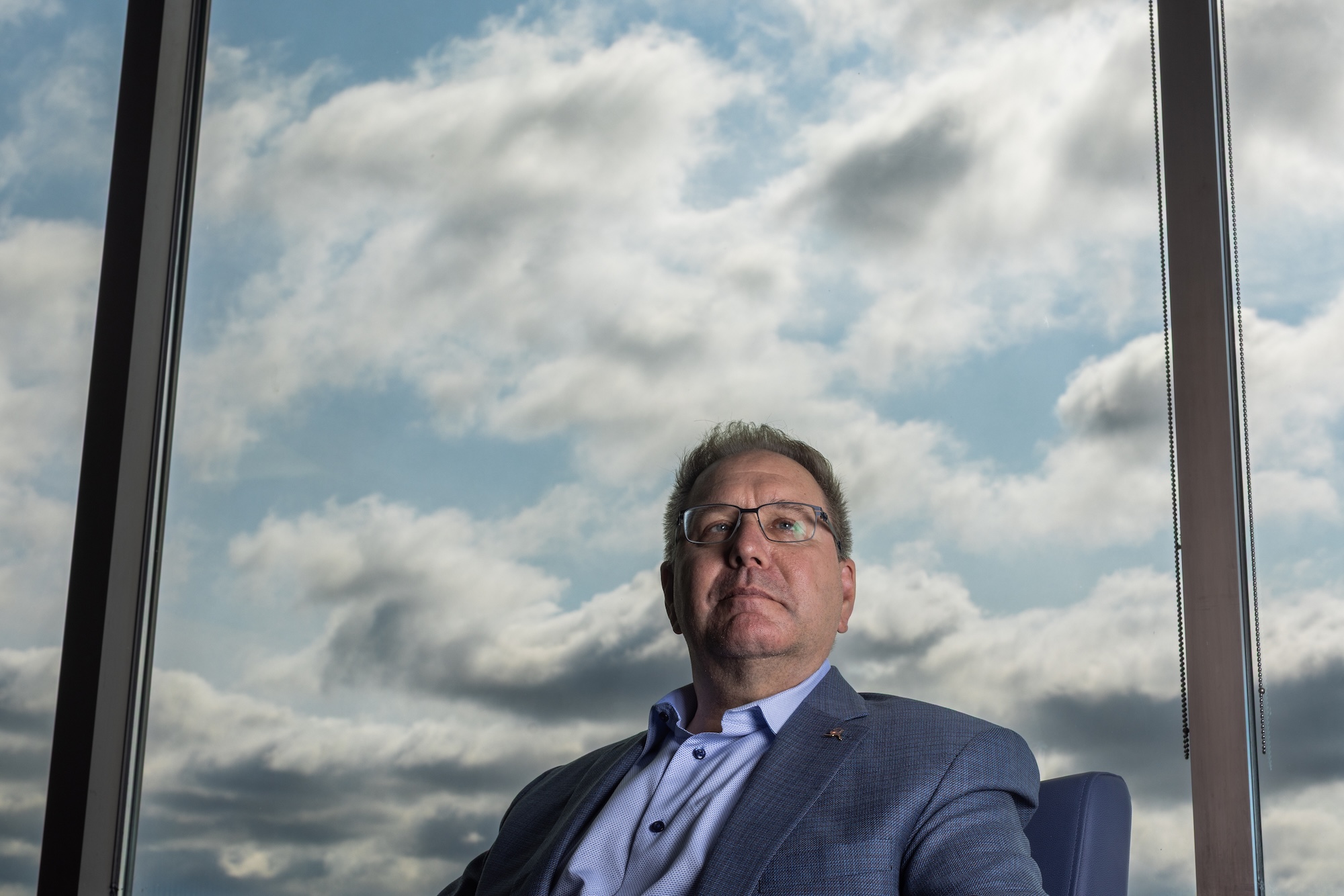 A man in a suit sits in front of a window showing a cloudy blue sky