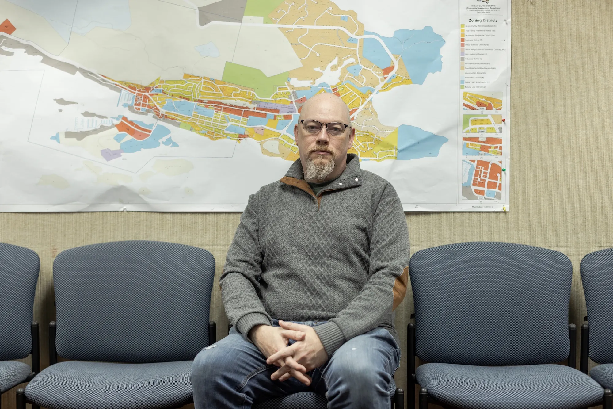 A man in glasses and a sweatshirt sits in a chair in front of a map