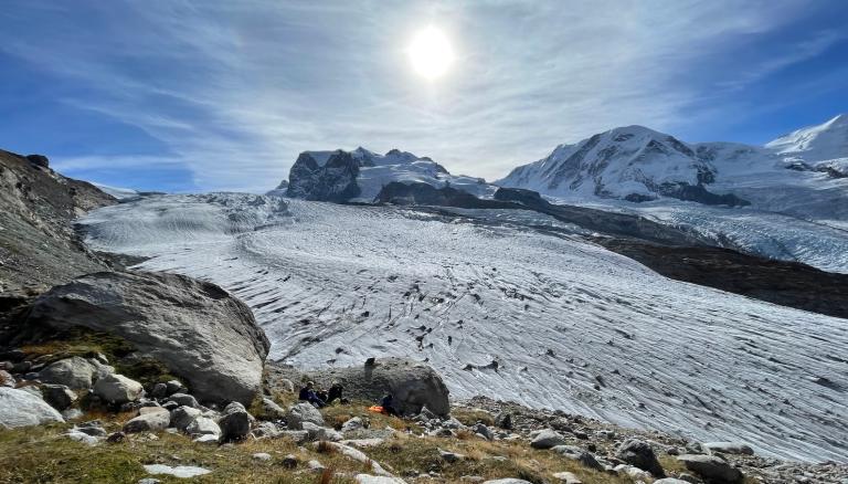 The sun shines over an icy mountain glacier, with a few researchers sitting on a grassy hill in the foreground