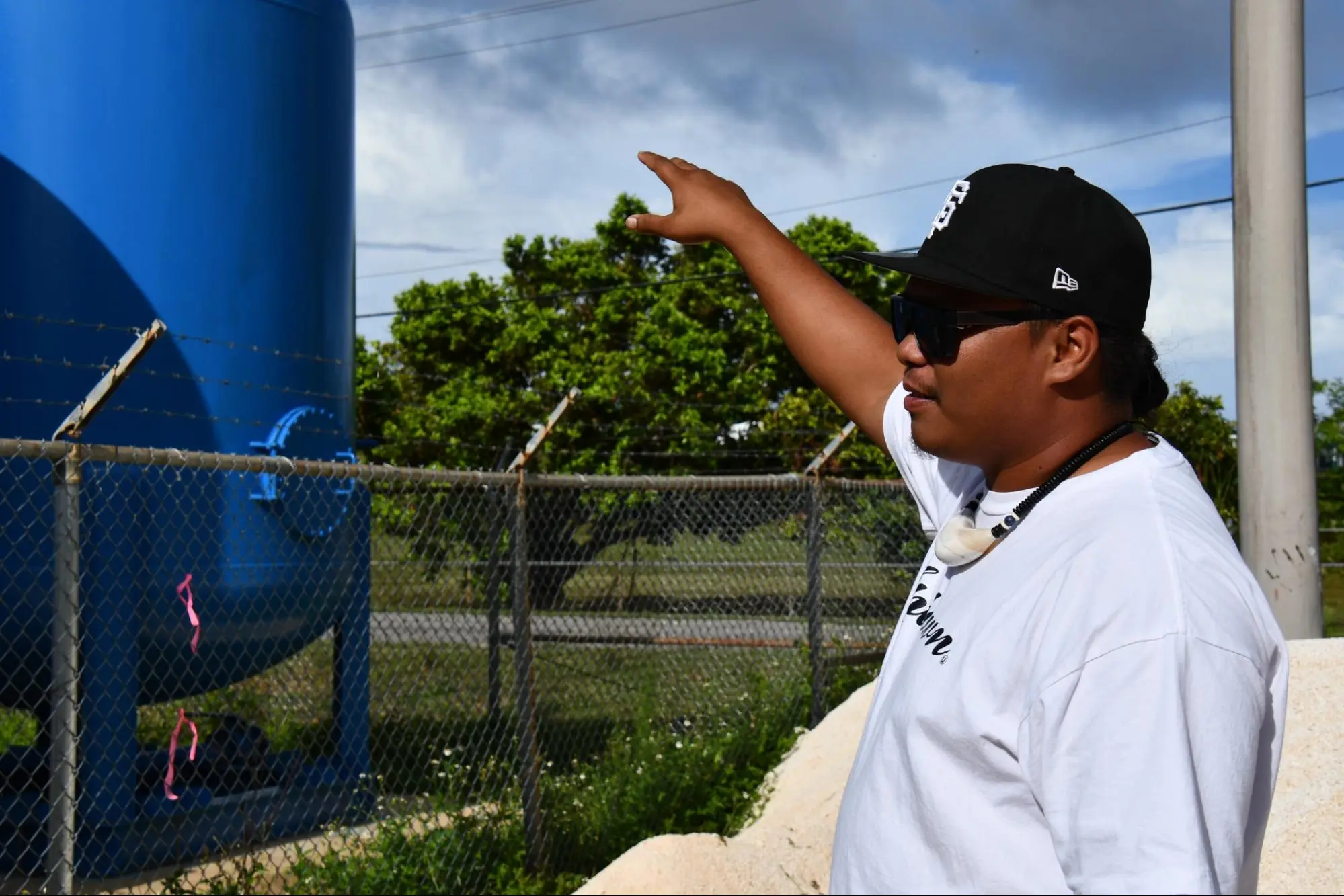 A man in a hat points at a building beyond a barb wire fence