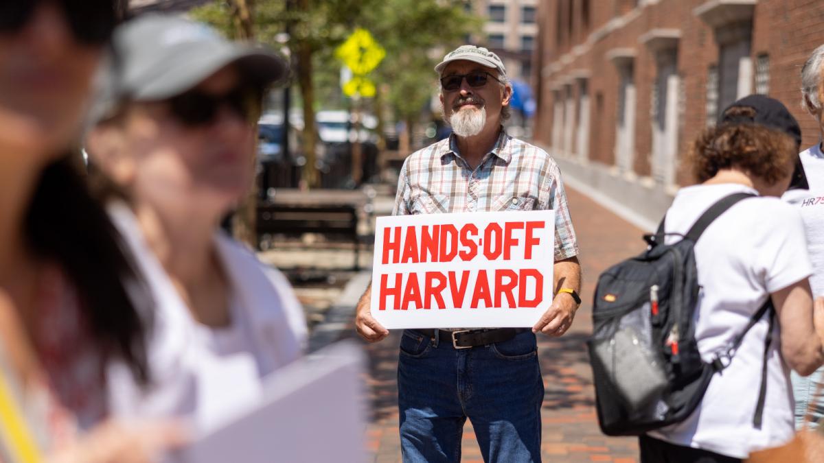 Photo of a man holding a sign that says hands off Harvard