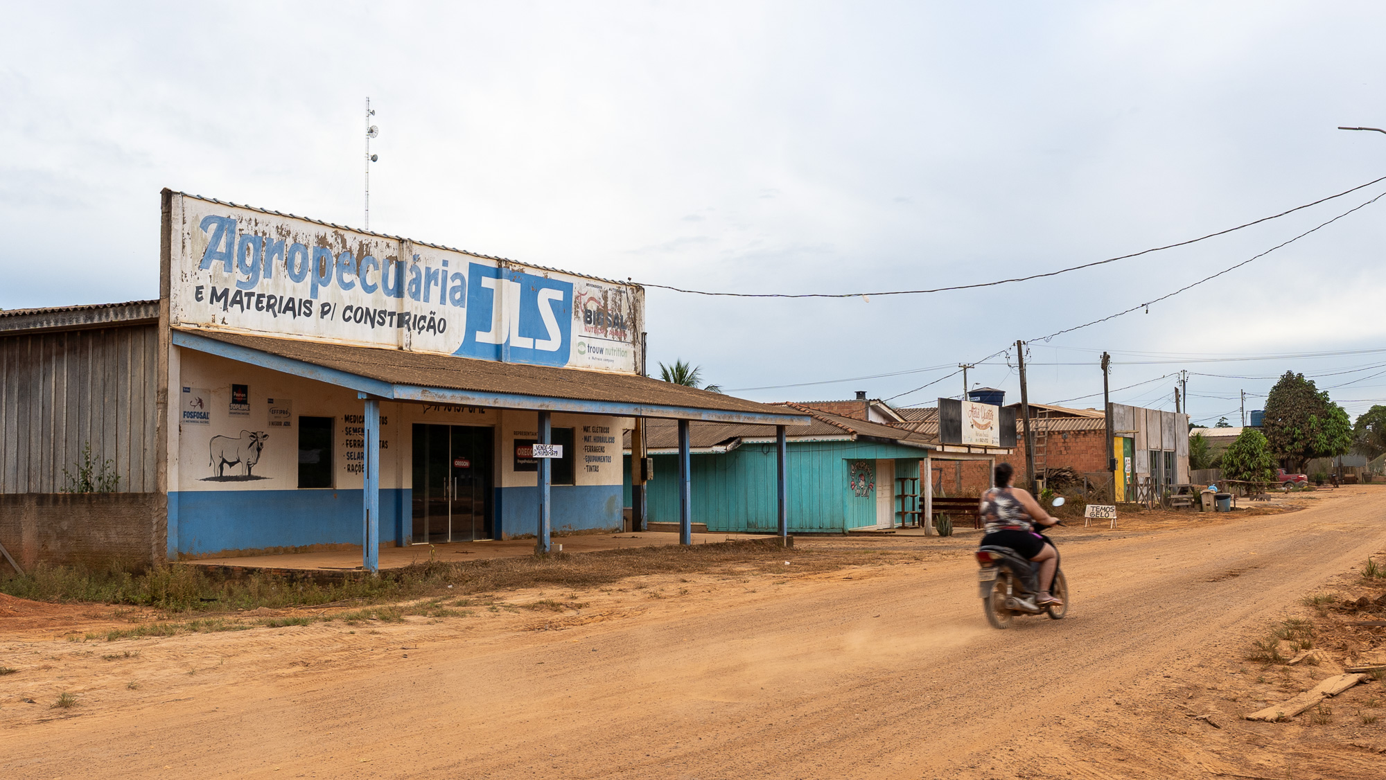 A dusty town with a business in Portuguese. A motorcycle drives by on an unpaved road
