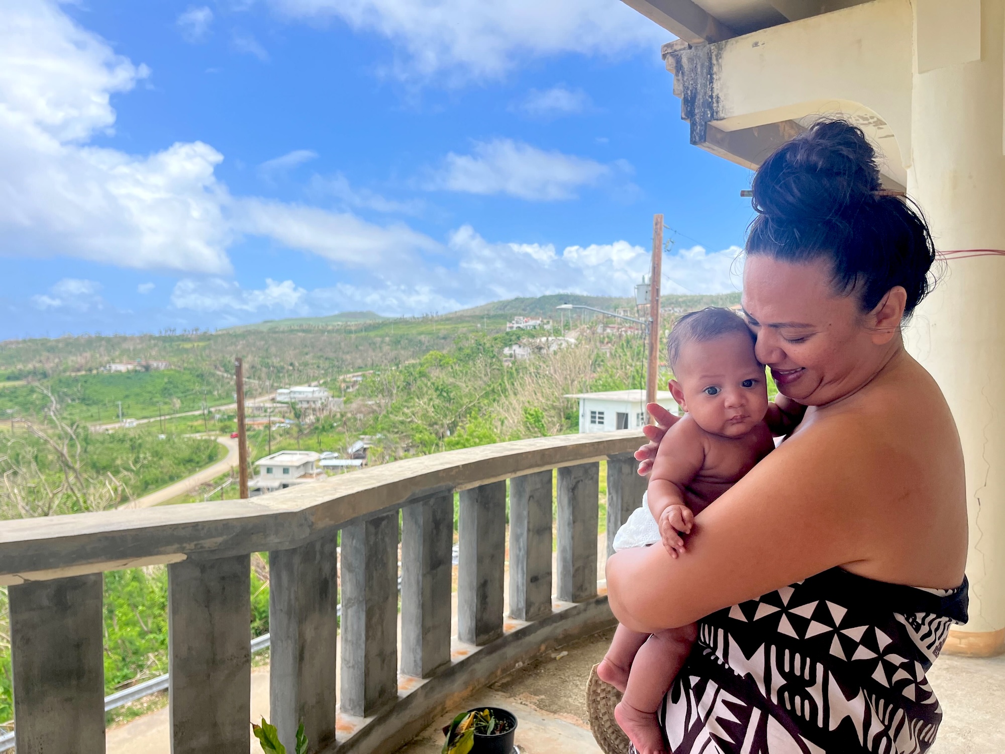 A woman holds a baby standing on a porch or balcony