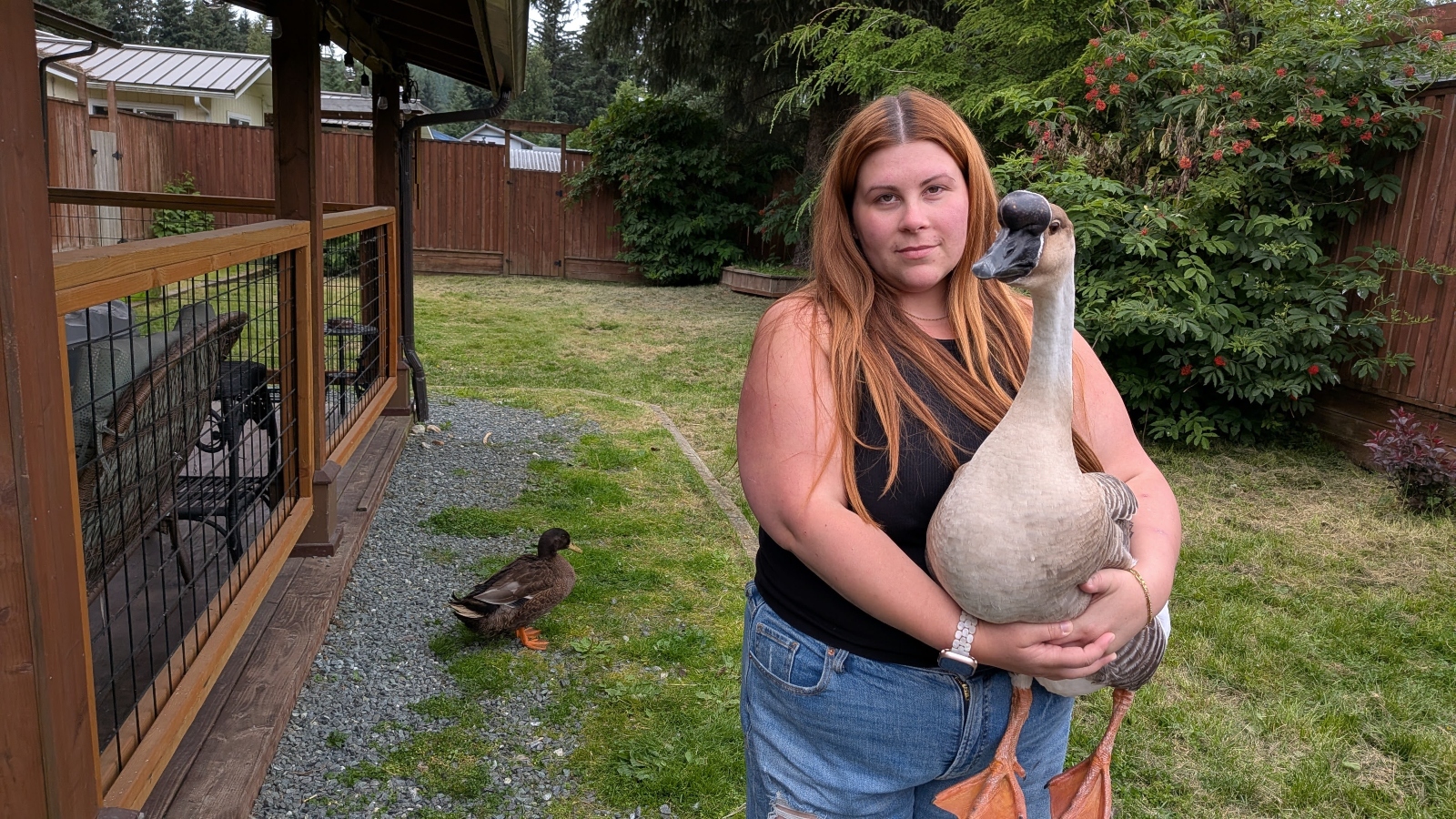 A woman with long hair looks into the camera while standing in a backyard holding a pet duck