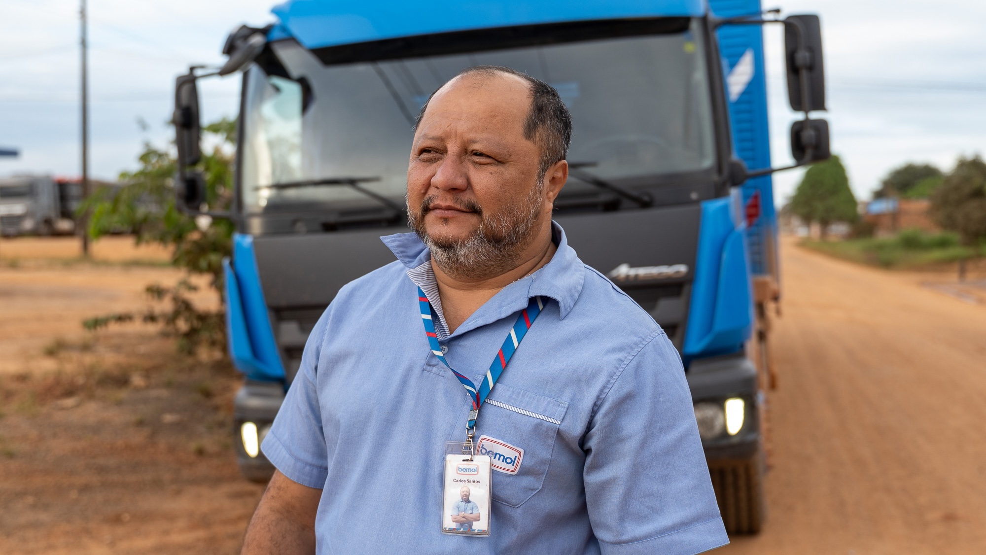 A man in a blue polo shirt and wearing an ID tag stands in front of a truck on an unpaved road