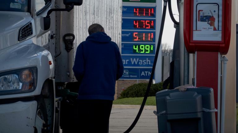A truck driver fills his big-rig with diesel fuel. A sign in the background indicates the price is four dollars and ninety-nine cents a gallon.