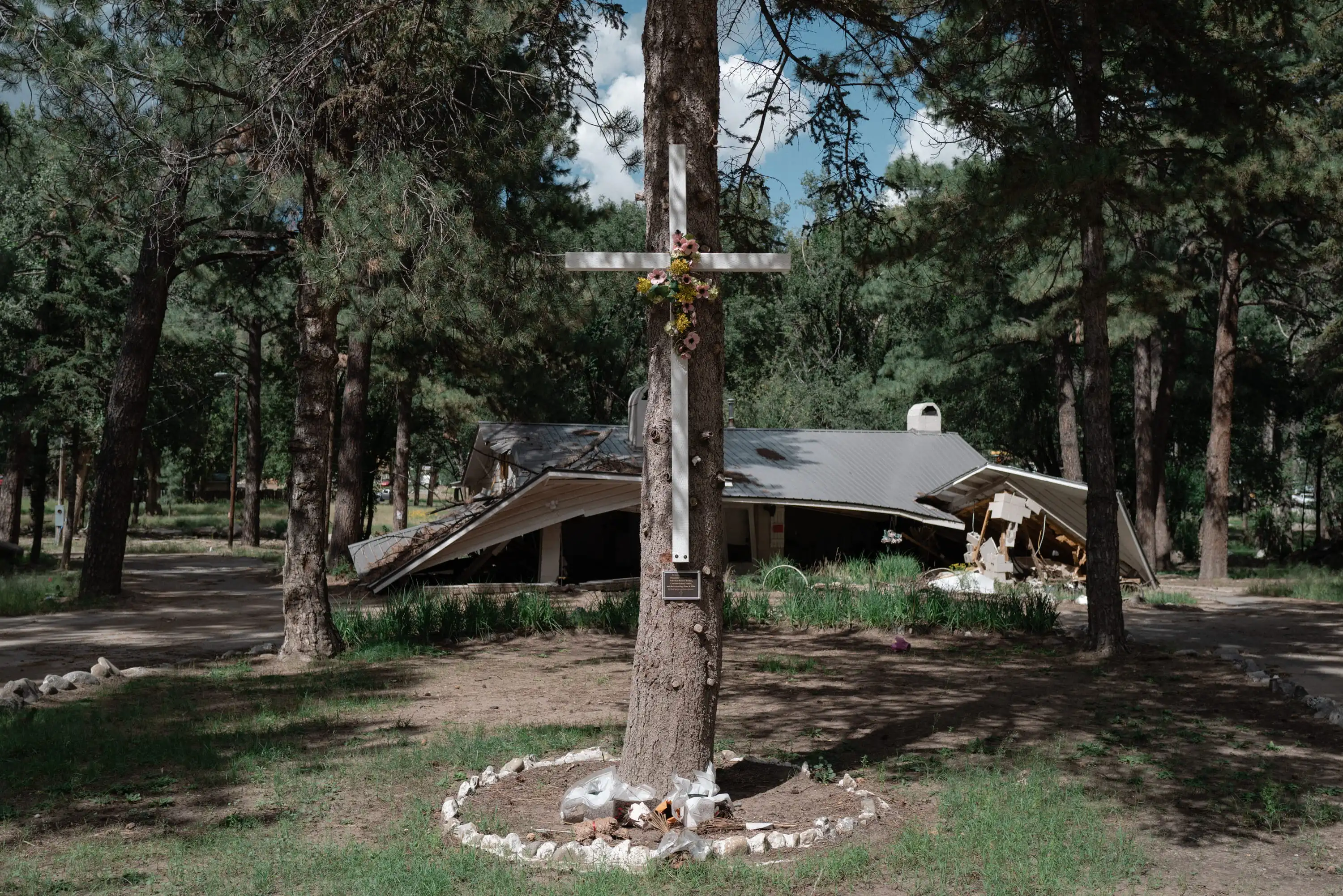 A wooden cross decorated with flowers stands attached to a tree in front of a collapsed building in a wooded area.
