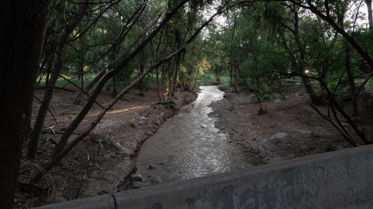 A shallow river, viewed from over a concrete barrier, flows through a dirt channel lined by trees and bare earth.