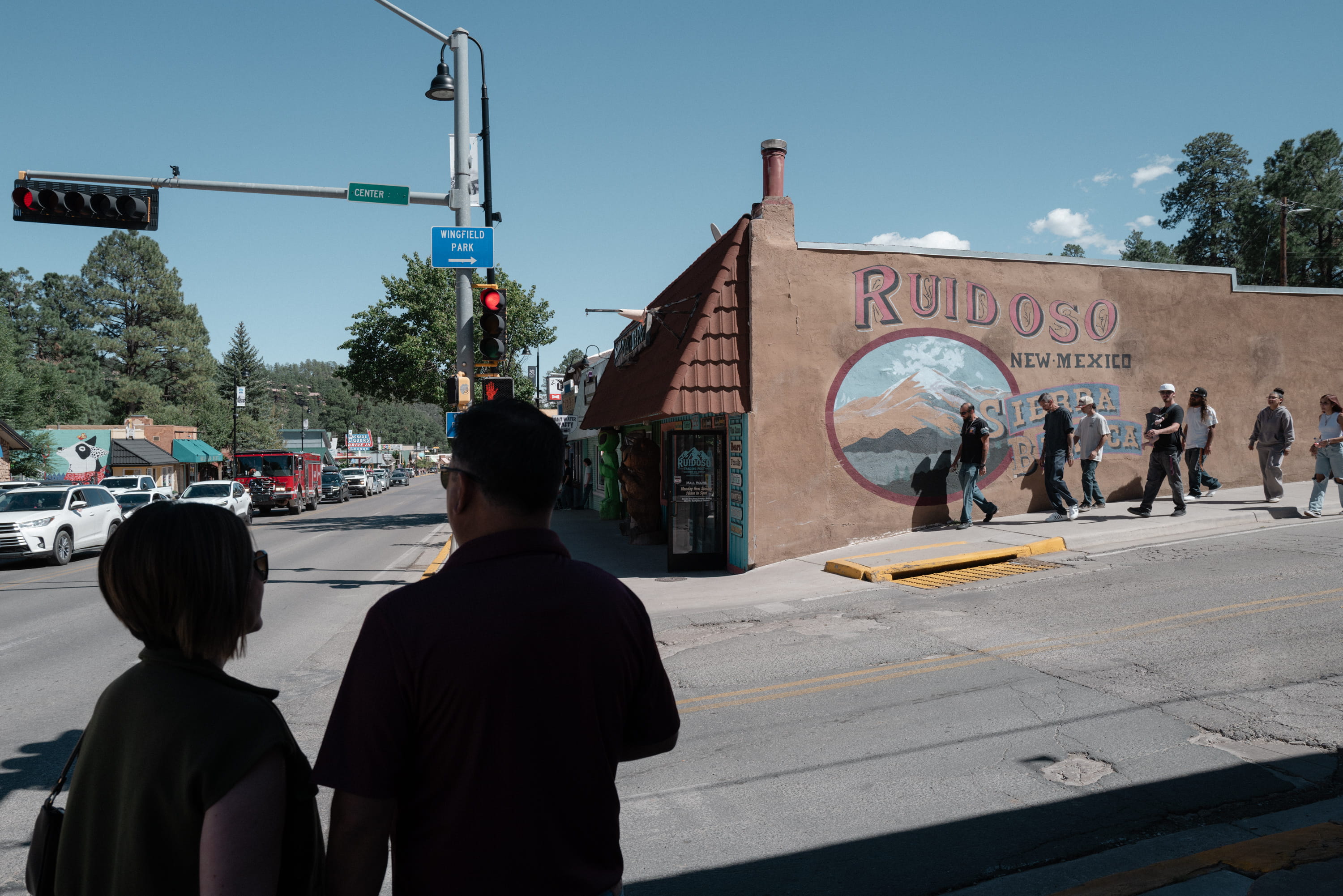 Pedestrians stand near a street corner in downtown Ruidoso, across from a large adobe wall featuring a welcome mural painted with the town's name and Sierra Blanca peak.