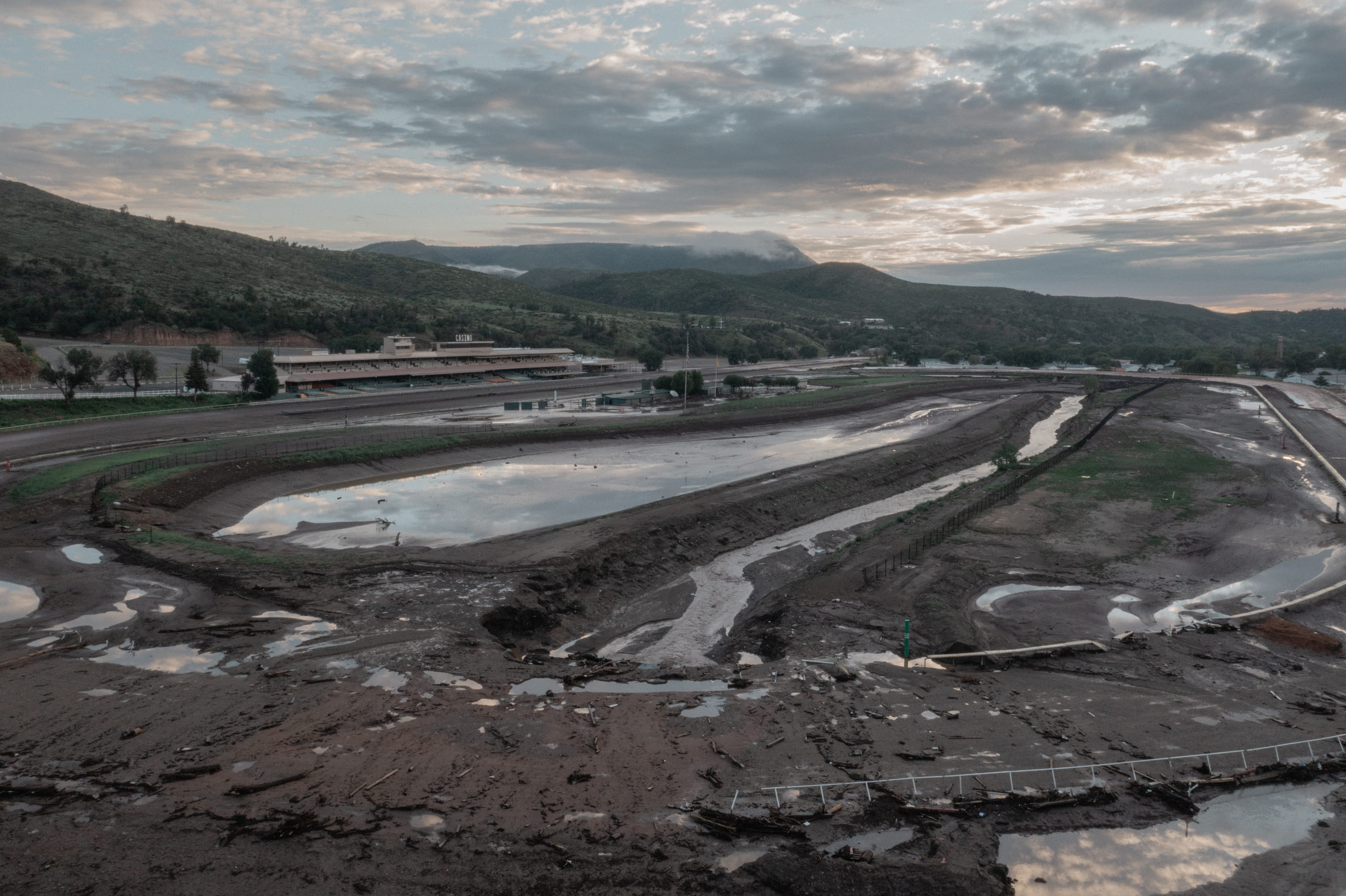 An elevated view shows a mud-caked, flooded horse-racing track surrounded by standing water and mountain ridges under a cloudy sky.