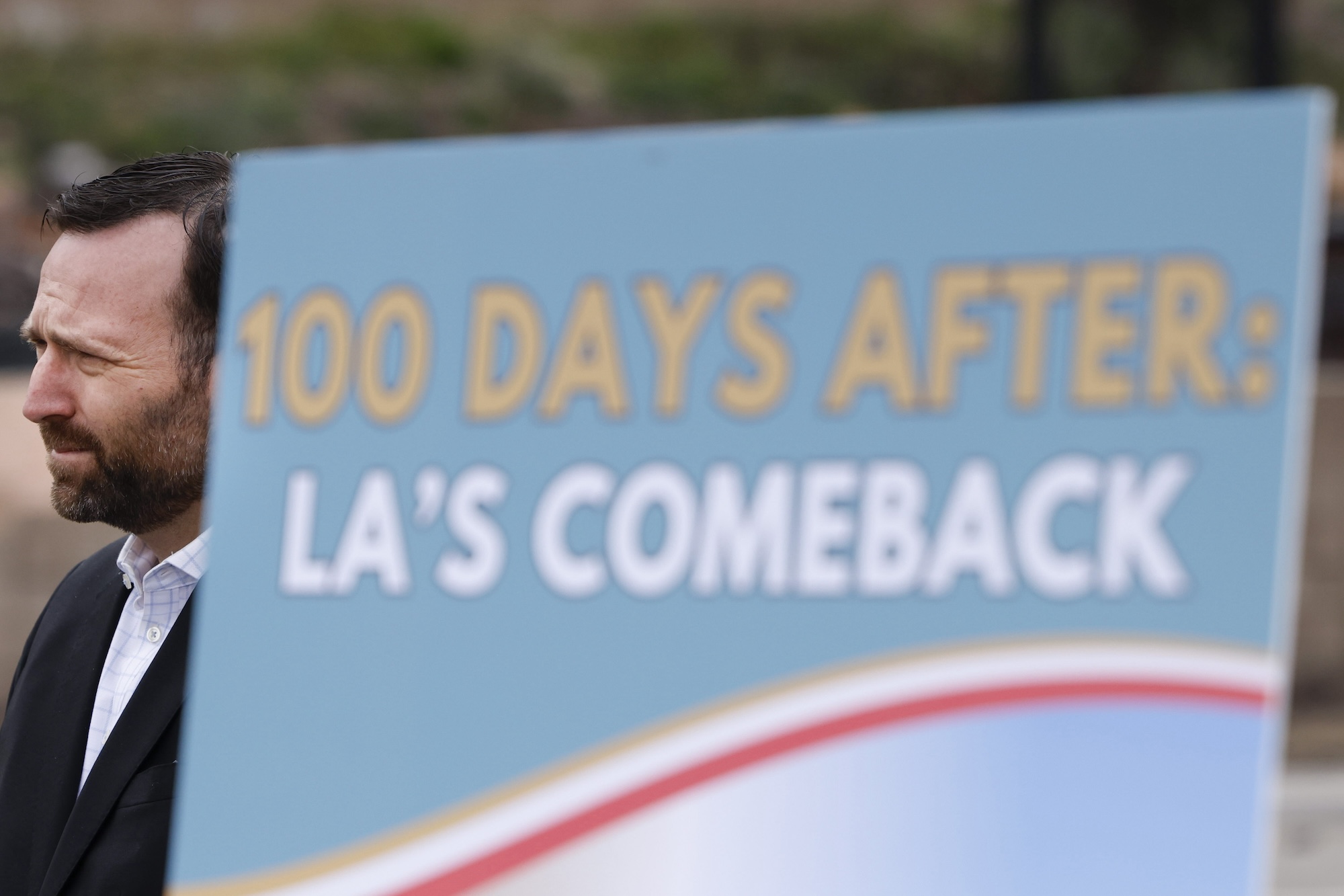 A man in a suit stands near a sign that says '100 days after: LA comeback'