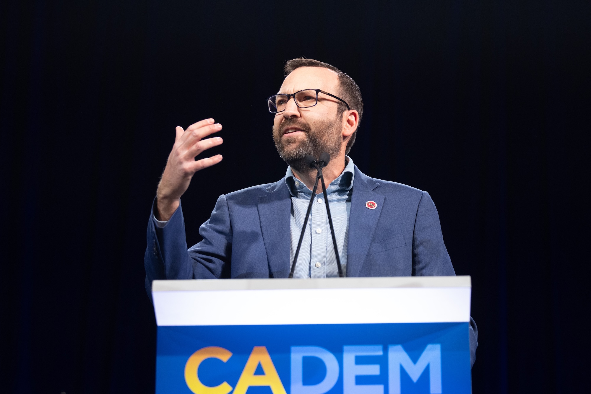 A man with glasses and a beard speaks behind a podium that says 'CADEM'