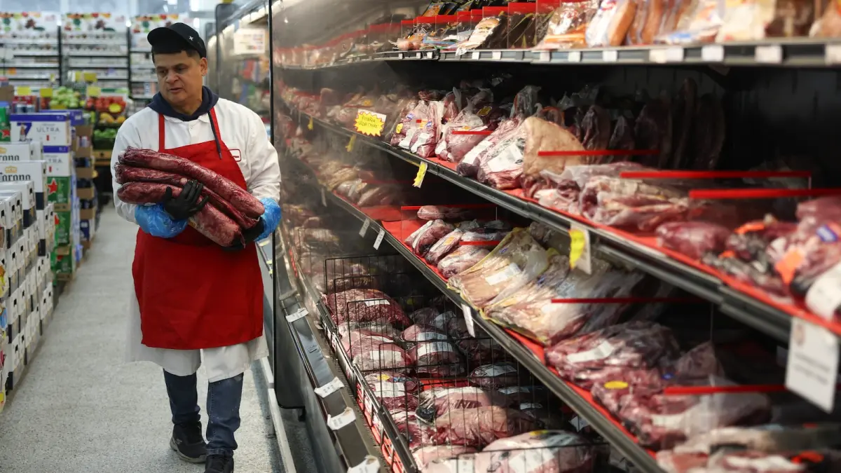 a grocery store butcher walks by the meat aisle holding three slabs of meat