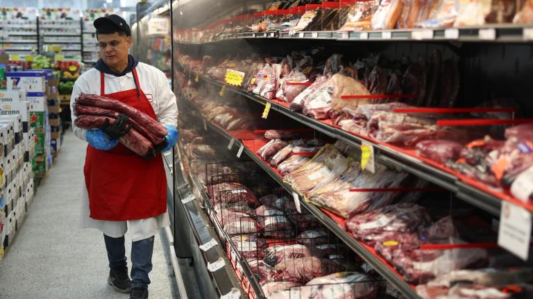 a grocery store butcher walks by the meat aisle holding three slabs of meat