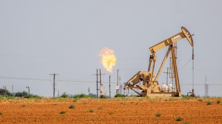 orange soil and a horizion line with trees cut across the bottom 1/4th of the image, a large piece of yellow machinery seen sitting on top of the horizon line, in the distance metal towers poke up out of the horizon line, one of them has a large flame on top