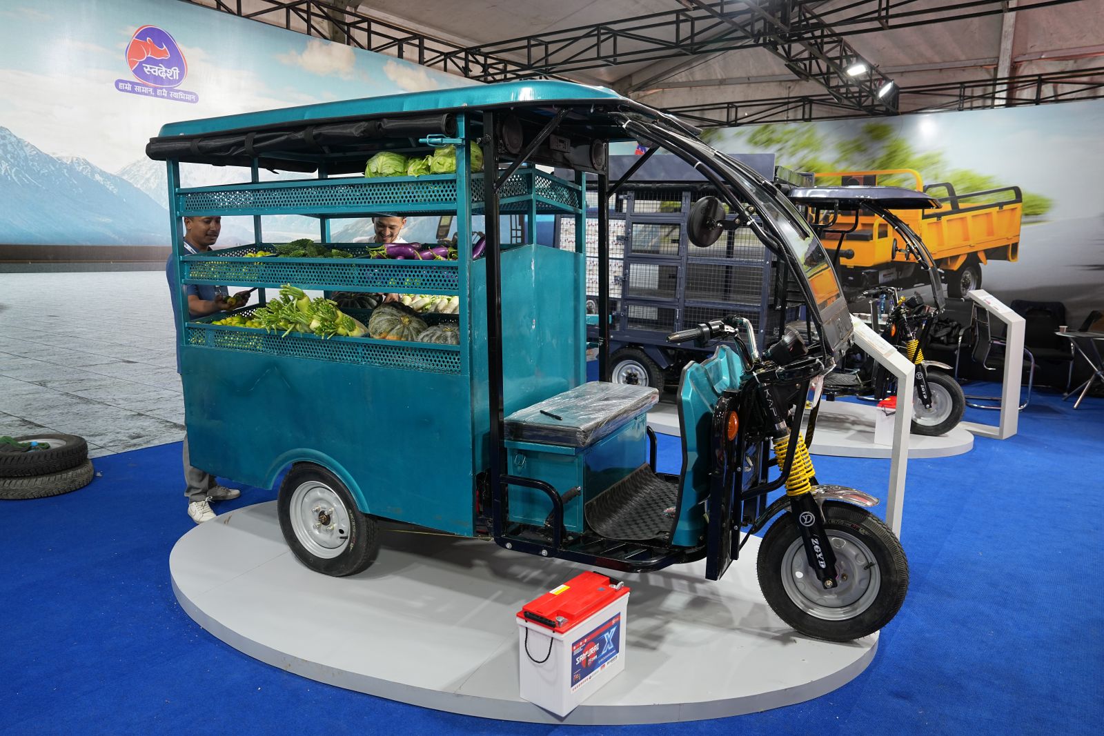 An electric tuktuk sits beside its battery at a trade show