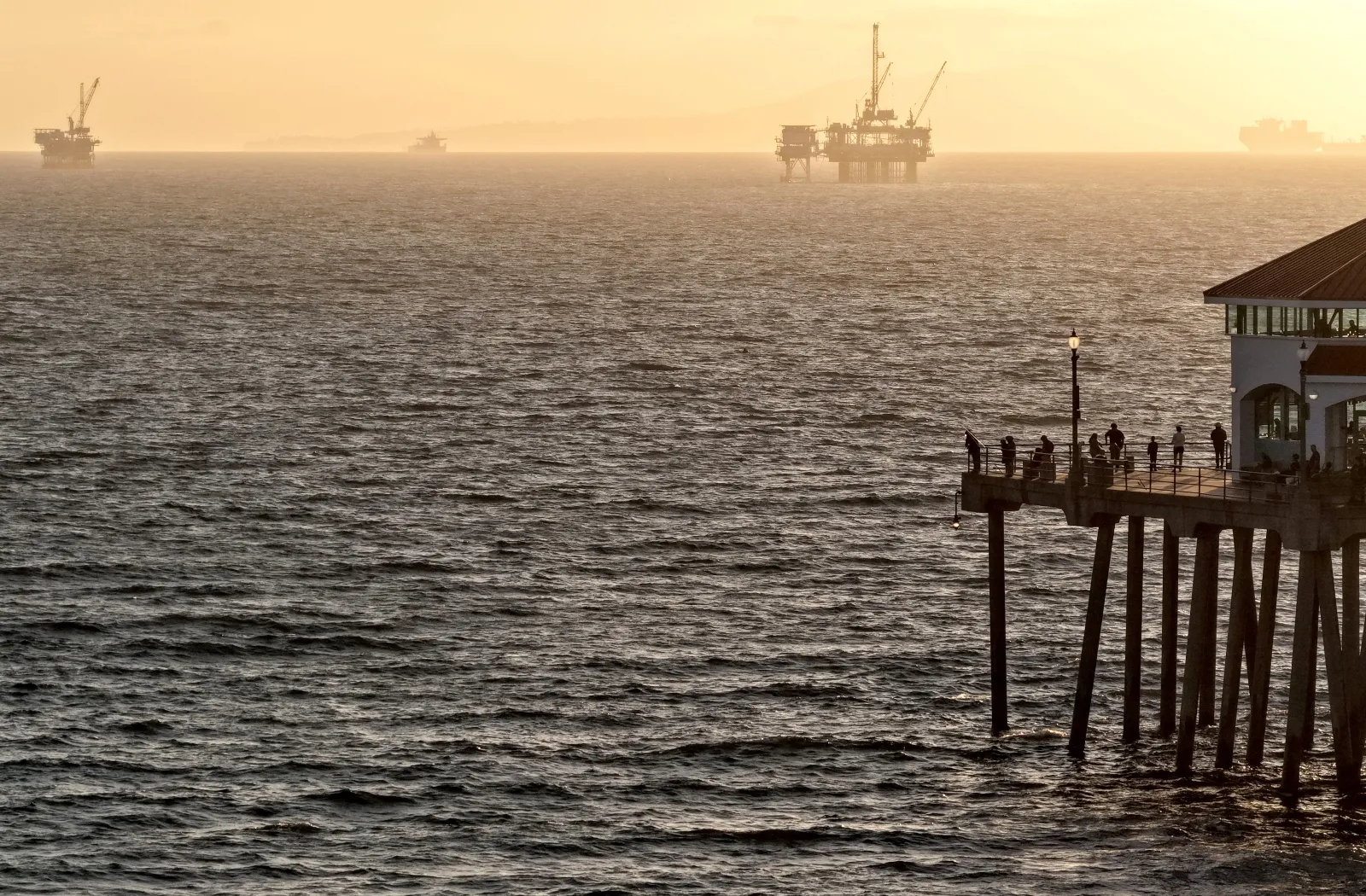 People stand above the Pacific Ocean on Huntington Beach pier, with the offshore oil and gas platform Eva in the distance. President Trump has sought to use his energy emergency declaration to enable more offshore oil drilling in the Gulf of Mexico.