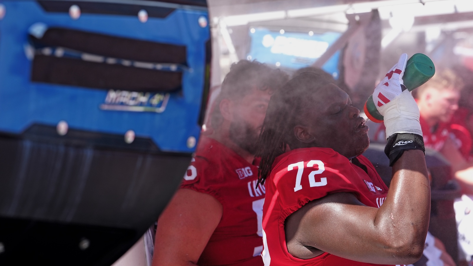 Two college football players sit in front of a large fan and sip water as they attempt to keep cool during a game.