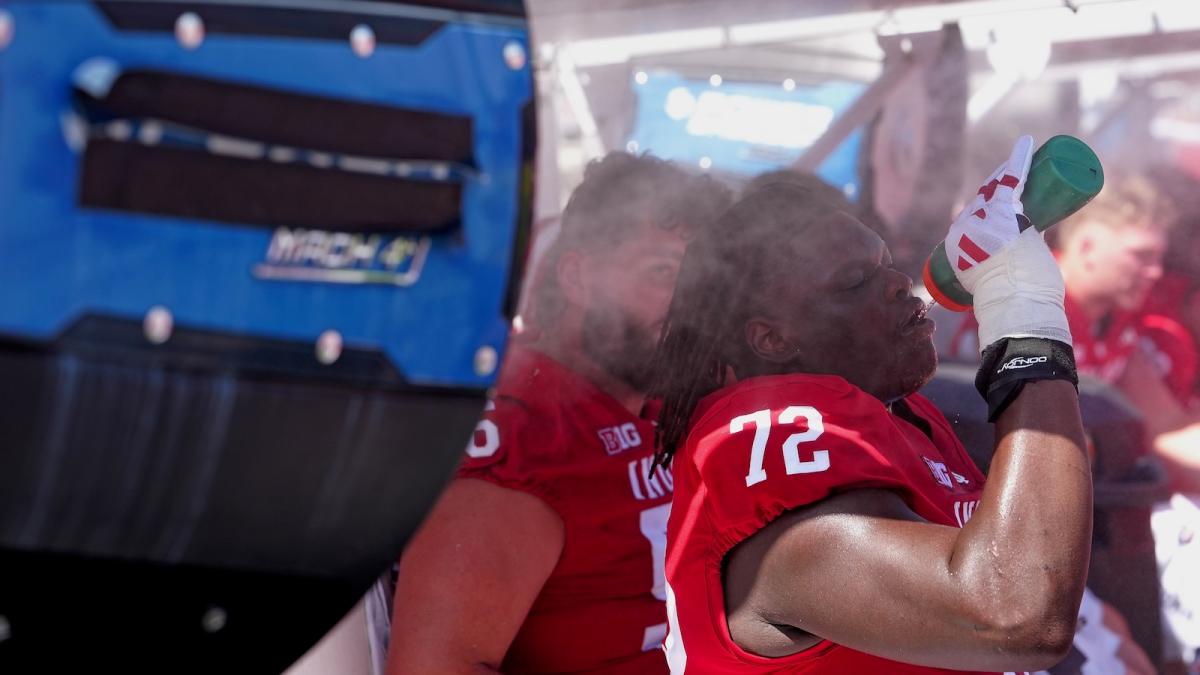 Two college football players sit in front of a large fan and sip water as they attempt to keep cool during a game.